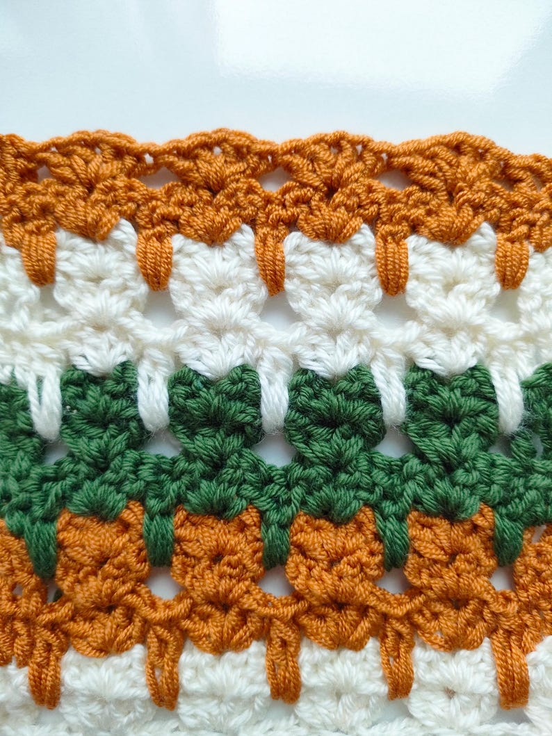 May include: Close-up of a crocheted fabric with alternating rows of orange, white, and green. The fabric has a textured, lacy pattern.