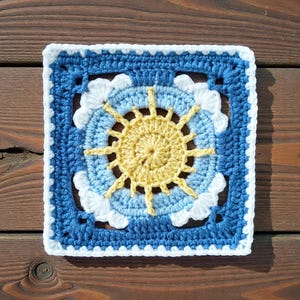May include: A square, handmade crochet piece featuring a sun design. The sun is yellow, surrounded by light blue and white accents, set against a dark blue background. The edges are finished with a white border.