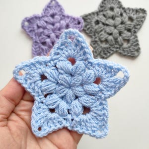 May include: Three crocheted star shapes in different colours: light blue, purple and grey. The stars are made with a textured stitch pattern.