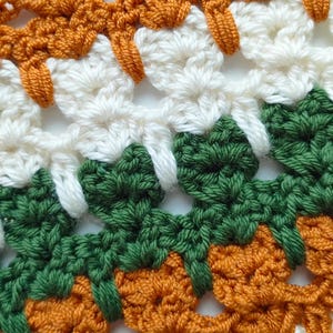 May include: Close-up of a crocheted fabric with a repeating pattern of orange, white, and green. The fabric is made with a variety of stitches and has a textured appearance.