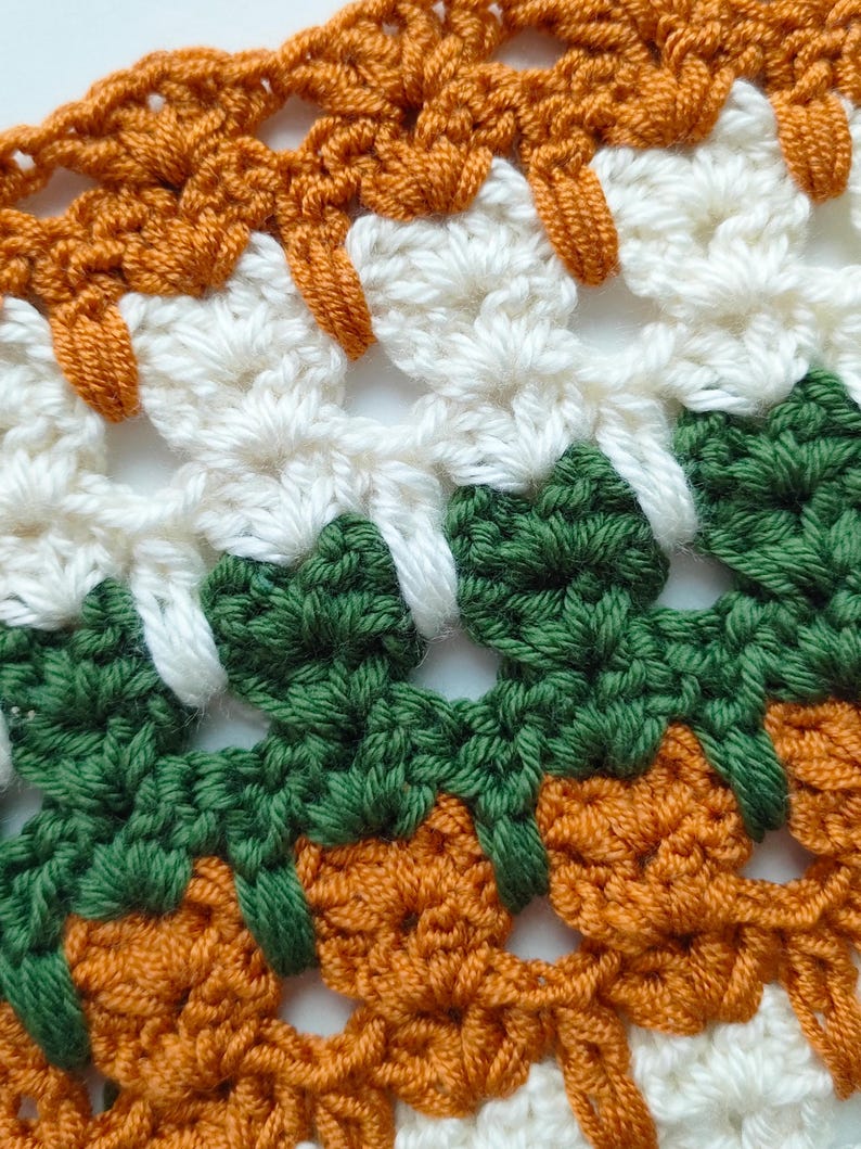 May include: Close-up of a crocheted fabric with a repeating pattern of orange, white, and green. The fabric is made with a variety of stitches and has a textured appearance.