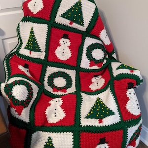 May include: A handmade crochet Christmas blanket. The blanket features a patchwork design with red, white, and green squares. Each square displays a Christmas tree, snowman, or wreath. The blanket is draped over a chair.