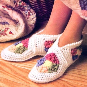 Può includere: Pantofole bianche all'uncinetto con disegni floreali colorati. Le pantofole presentano una punta quadrata e un design a taglio basso, con un mix di filato rosa, verde, giallo e blu. Le pantofole sono su un pavimento in legno.