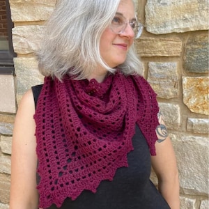 May include: A woman wearing a burgundy crocheted shawl with a scalloped edge. The shawl is made with a delicate stitch pattern and is draped around her neck.