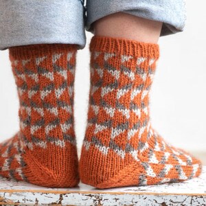 Puede incluir: Un par de calcetines de punto con un patr&oacute;n geom&eacute;trico. Los calcetines son principalmente de color naranja con un dise&ntilde;o repetido en gris y blanco. Los calcetines son visibles desde los tobillos hacia abajo, con los pu&ntilde;os de unos vaqueros azules encima.