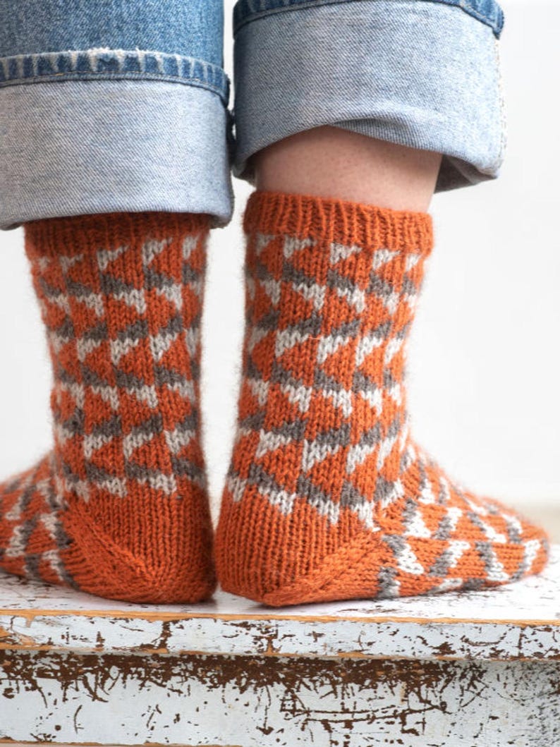Puede incluir: Un par de calcetines de punto con un patr&oacute;n geom&eacute;trico. Los calcetines son principalmente de color naranja con un dise&ntilde;o repetido en gris y blanco. Los calcetines son visibles desde los tobillos hacia abajo, con los pu&ntilde;os de unos vaqueros azules encima.
