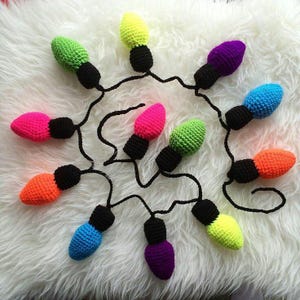 May include: A string of colorful, crocheted Christmas lights. The lights are in various colors, including pink, green, orange, blue, purple, and yellow. Each light has a black base and is connected by a black wire. The lights are arranged on a white, fluffy surface.