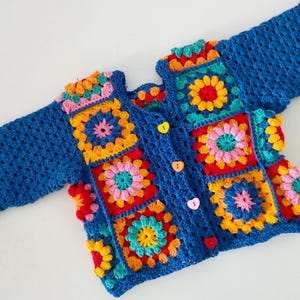 Puede incluir: Un cárdigan de ganchillo azul con cuadrados florales multicolores. El cárdigan tiene una parte delantera abotonada con botones en forma de corazón y mangas largas. Los cuadrados son en tonos de rojo, amarillo, rosa y naranja.