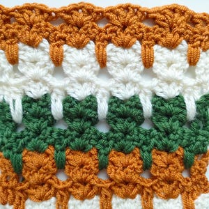 Op de afbeelding: Een close-up van een gehaakt stof met een herhalend patroon van oranje, wit en groen. De stof is gemaakt met verschillende steken en heeft een textuur.