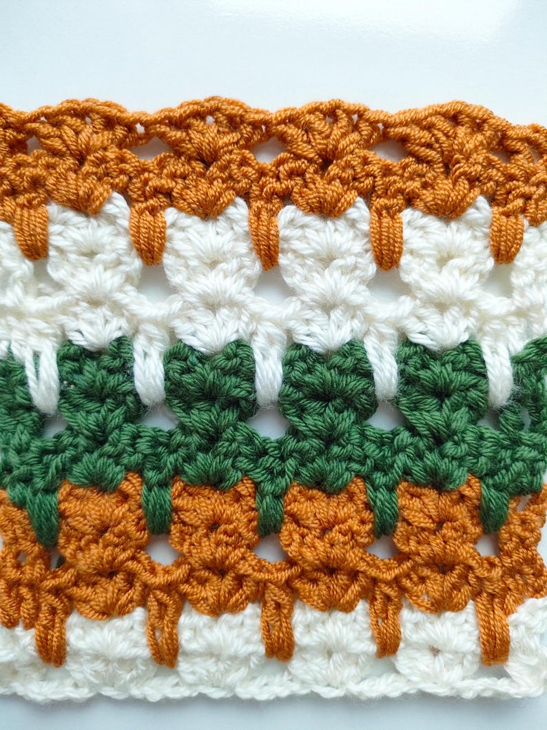 May include: A close-up of a crocheted fabric with a repeating pattern of orange, white, and green. The fabric is made with a variety of stitches and has a textured look.