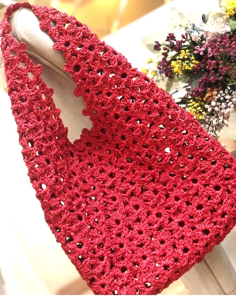 May include: A handmade red crochet tote bag with a floral pattern. The bag features a sturdy handle and a spacious interior. The crochet design is intricate, with a repeating flower motif. The bag is made of a soft, durable material, perfect for carrying everyday items.