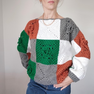 May include: A crocheted sweater with a patchwork design in shades of white, green, grey, orange, and brown. The sweater features a large floral motif in the centre.