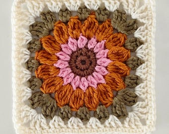 May include: A square, crocheted granny square with a sunflower design. The centre is brown, surrounded by pink petals, then orange petals, and olive green leaves. The border is cream-coloured.