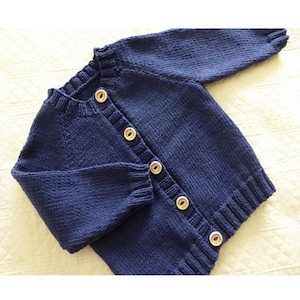 May include: A navy blue knitted cardigan with a round neckline and long sleeves. The cardigan has a button-up front with six round, light-coloured buttons. The knitwear has a ribbed hem and cuffs, and is likely made of wool or a similar material.