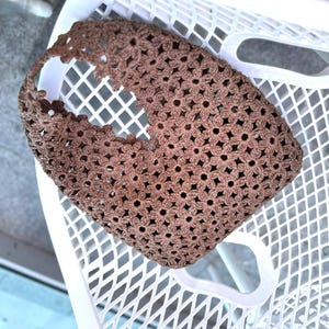 Puede incluir: Un bolso de mano marrón tejido a ganchillo con un patrón floral. El bolso tiene un asa y está hecho de un material tejido. El bolso está sobre una silla de plástico blanca.