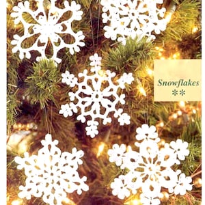 以下が含まれることがあります： クリスマスツリーに吊るされた、白の編み物で作られた雪の結晶のオーナメントが5つ。雪の結晶は複雑な透かし彫りのデザインで、細い糸で吊るされています。「Snowflakes」の文字が小さな薄緑色の箱に入っています。