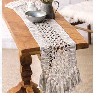 Op de afbeelding: Een lichtgrijze gehaakte tafelloper met een geometrisch openwerkontwerp en franjekwastjes. De loper wordt getoond op een houten tafel, waarbij de textuur en decoratieve aantrekkingskracht worden getoond. Een metalen kom en vaas staan ook op de tafel.