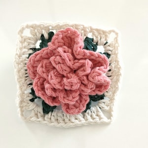 Op de afbeelding: Een gehaakt vierkant met een roze bloem in het midden. Het vierkant is gemaakt van wit, groen en roze garen.