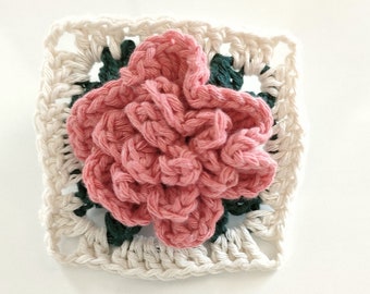 May include: A crocheted square with a pink flower in the center. The square is made with white, green, and pink yarn.