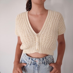 Peut inclure: Cardigan en crochet crème à manches courtes, col en V et fermeture boutonnée. Le haut est associé à un jean en denim délavé. Le motif en crochet est visible, et les boutons correspondent à la couleur du cardigan.