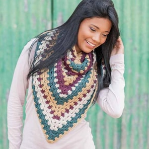 May include: A colorful, crocheted shawl in a triangular shape. The shawl features a granny square design with a palette of teal, purple, cream, and tan. The person is wearing a light pink long-sleeved shirt.