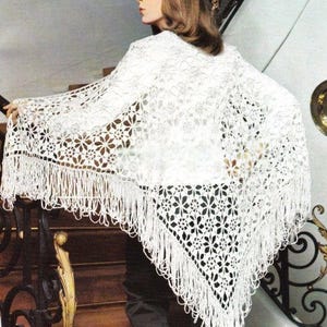 Puede incluir: Un chal blanco de crochet calado con un patrón floral y flecos. El chal está drapeado sobre los hombros. El diseño intrincado y la textura delicada crean una estética vintage y hecha a mano.