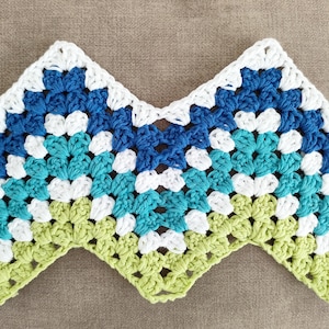 May include: A handmade crochet piece in a chevron pattern. The piece features a color gradient of white, blue, and green. The crochet work is visible, with each stitch clearly defined. The item is likely a decorative element or part of a larger textile project.