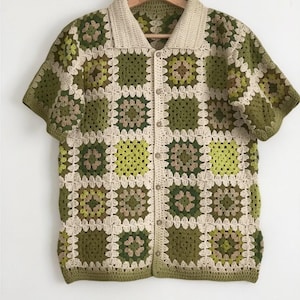 May include: A short-sleeved crochet shirt with a button-down front. The shirt has a patchwork design of green, olive, and beige squares. The collar and button placket are beige. The shirt is displayed on a wooden hanger.