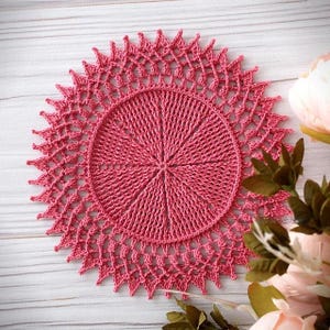 May include: A circular, dusty rose-colored crochet doily featuring a dense, radiating center pattern that transitions into an intricate, open-work scalloped border with pointed edges, creating a sunburst effect in the overall design.