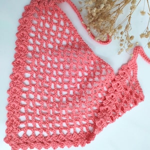 May include: A coral-colored triangular crochet bandana featuring an openwork diamond mesh pattern and a decorative scalloped edge. The accessory includes two thin crocheted ties at the top corners for fastening, showcasing a detailed yarn texture.