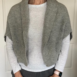 May include: A gray knitted shawl with a textured pattern. The shawl is draped over a light gray long-sleeved shirt. The shawl has a wide collar and short sleeves. The overall look is casual and comfortable.