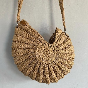 May include: A natural-toned raffia shoulder bag. The bag features a rounded design with a wide opening and a braided strap. The bag is approximately 30 cm wide and 25 cm tall, ideal for carrying essentials.