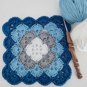 Puede incluir: Un cuadrado de ganchillo hecho a mano en tonos azules, grises y blancos. Presenta un borde festoneado y un patrón floral en el centro. Un ganchillo de madera y dos ovillos de lana, uno azul y otro blanco, están junto al cuadrado.