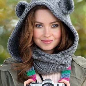 May include: A gray knitted hood with bear ears and a matching scarf with colorful stripes. The hood is worn by a person holding a Canon camera. The background is blurred with shades of green and brown.