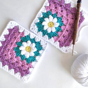 Puede incluir: Dos cuadrados de granny crochet con una flor de margarita blanca en el centro. Los cuadrados están hechos con hilo rosa, blanco y turquesa. También hay una bola de hilo blanco y un gancho de crochet de madera en la imagen.
