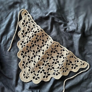 Pode incluir: Uma bandana triangular de crochê na cor bege claro com um intrincado padrão de conchas vazadas, bordas festonadas e finas tiras de crochê nos dois cantos superiores para prender a peça ao redor da cabeça ou do pescoço.
