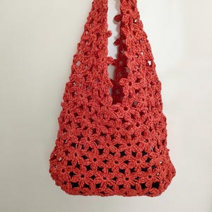 May include: A coral-colored crochet tote bag, featuring a floral pattern. The bag has two handles and a spacious interior. The bag is made of a soft, textured yarn and is suitable for carrying various items.