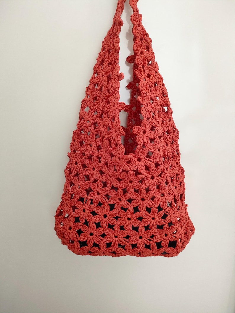 May include: A coral-colored crochet tote bag, featuring a floral pattern. The bag has two handles and a spacious interior. The bag is made of a soft, textured yarn and is suitable for carrying various items.