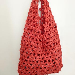 May include: A coral-colored crochet tote bag. The bag is made of a floral pattern and has a handle for carrying. The bag is hanging against a white background.