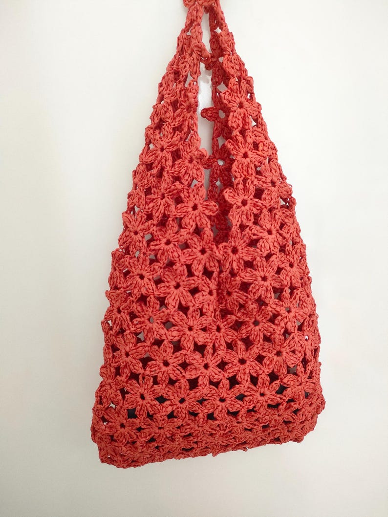 May include: A coral-colored crochet tote bag. The bag is made of a floral pattern and has a handle for carrying. The bag is hanging against a white background.