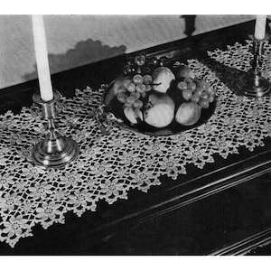 Puede incluir: Una foto en blanco y negro de un camino de mesa de ganchillo con un delicado patrón floral. El camino de mesa está colocado sobre una mesa de madera oscura con un cuenco de fruta y dos candelabros.