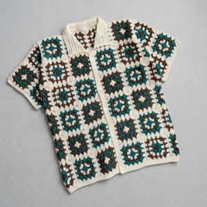 Può includere: Una camicia a maniche corte con bottoni e motivo all'uncinetto. La camicia presenta una base color crema con motivi quadrati marroni e verde acqua. Il colletto e l'abbottonatura sono color crema.