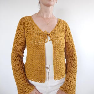 May include: Mustard yellow crochet cardigan with long sleeves and a V-neck. The open-front design features a tie closure at the neckline. The cardigan is made of a lightweight, open-weave material, perfect for layering.