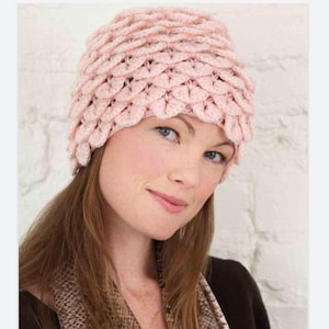 May include: A light pink crocheted hat with a textured, scale-like pattern. The hat is a soft, pastel color and appears to be handmade. The hat is a winter accessory.