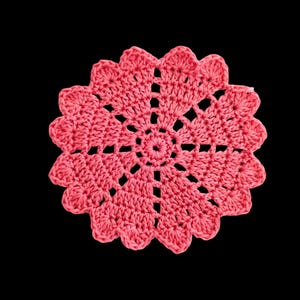 May include: A handmade, coral-coloured crochet doily with a floral design. The doily features a central circular pattern surrounded by openwork and scalloped edges. The craftwork is set against a black background.