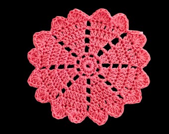 Peut inclure: Un napperon au crochet fait à la main, de couleur corail, avec un motif floral. Le napperon présente un motif circulaire central entouré de jours et de bords festonnés. Le travail artisanal est posé sur un fond noir.