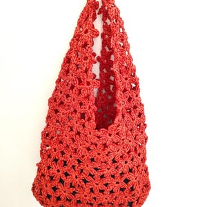 May include: A red crocheted bag with a floral pattern. The bag is made of a woven material and has a handle. The bag is hanging against a white background. The bag's interior is dark.