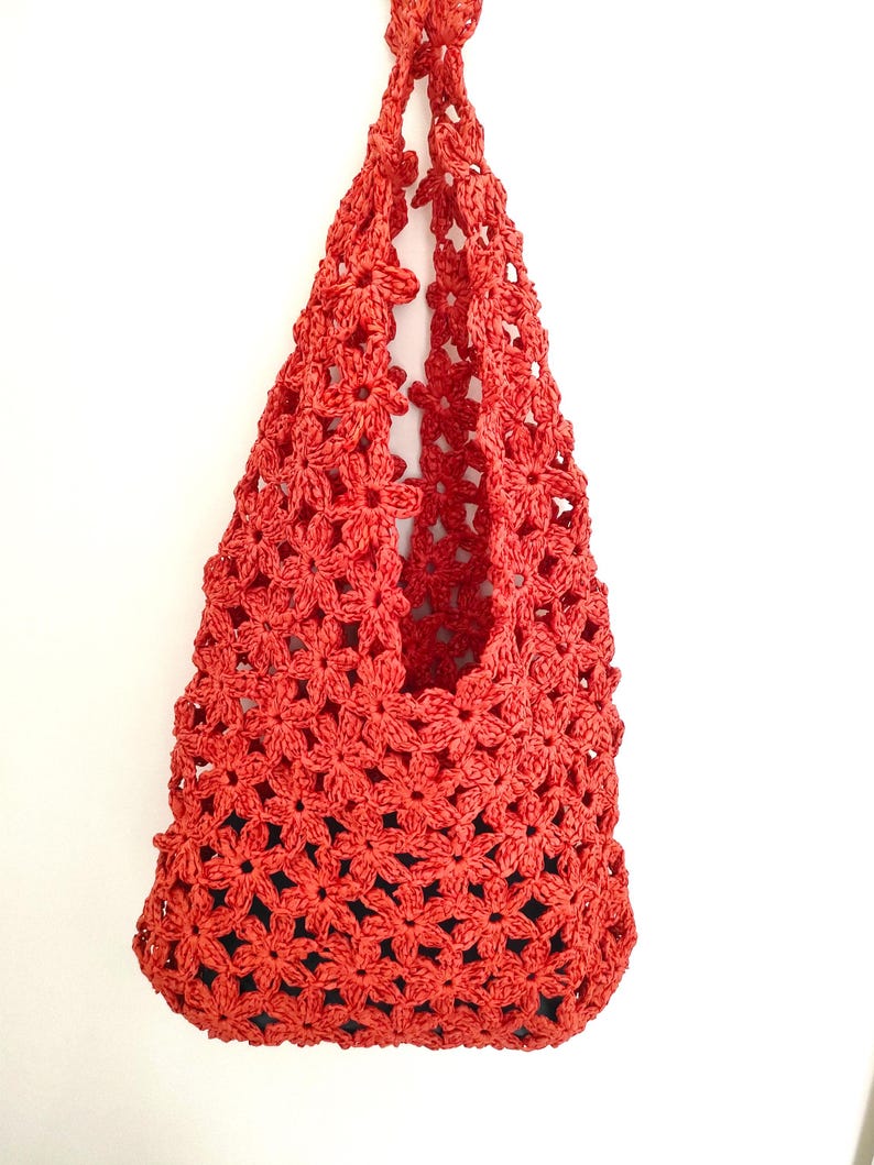 May include: A red crocheted bag with a floral pattern. The bag is made of a woven material and has a handle. The bag is hanging against a white background. The bag's interior is dark.