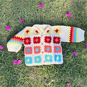 Puede incluir: Un cárdigan de crochet blanco con un patrón geométrico colorido. El cárdigan tiene cierre de botón y mangas largas.