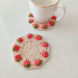 Puede incluir: Dos posavasos de ganchillo con un diseño de fresa. Los posavasos son de color beige con fresas rosas y verdes. Una taza de café blanca está sobre uno de los posavasos.
